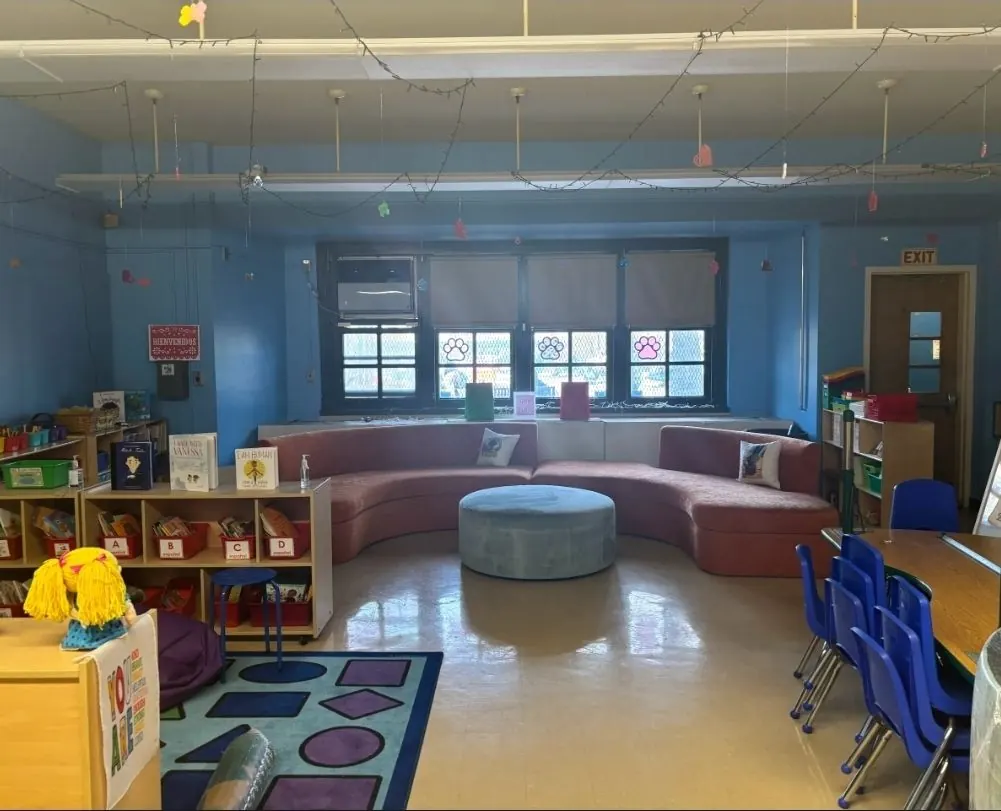 A bright blue classroom with a curved pink sofa, a round gray ottoman, book bins, and small blue chairs