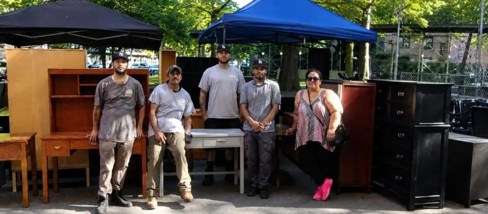 Four movers and a volunteer standing among donated dressers and hutches under pop-up tents