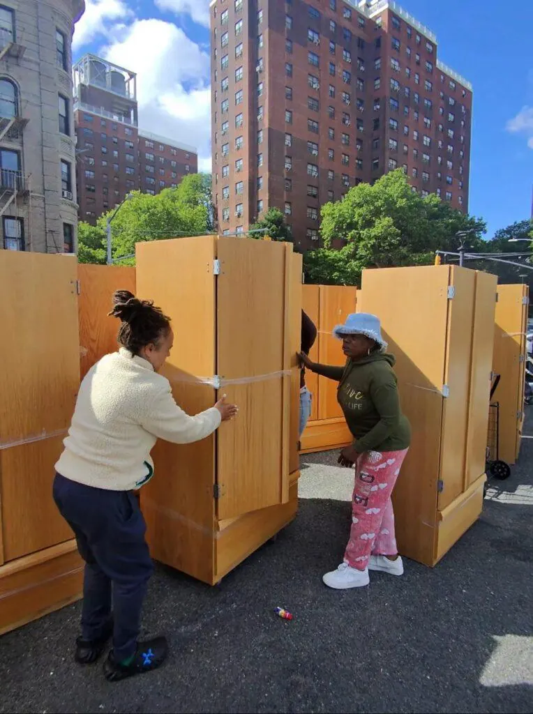 Two residents unwrapping donated oak wardrobes in a parking lot beneath NYCHA towers